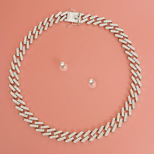 Overhead flat lay of a wide silver tone fully pave clear rhinestone square Cuban chain link necklace arranged in a circle on a coral pink surface. A square push-lock box clasp covered in clear rhinestones is visible at the top. Two small clear rhinestone round stud earrings with clear rubber butterfly post backings are displayed inside the necklace loop.