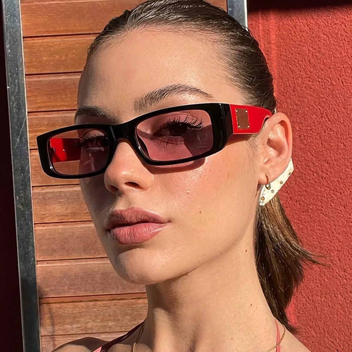 Woman wearing black rectangular sunglasses with red temples, pink-toned lenses, and gold-tone accents against wood panels and a red wall