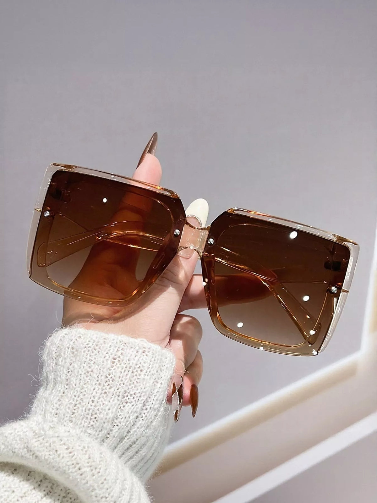 Caramel translucent square sunglasses with brown gradient lenses, held by a manicured hand in a white knit sleeve