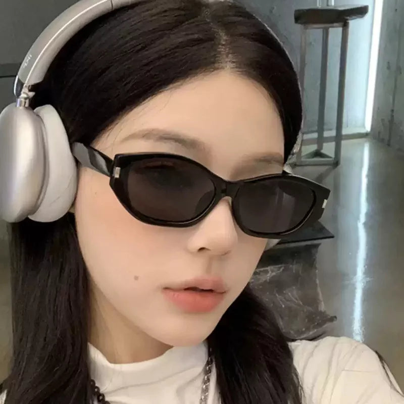 Woman wearing black angular cat-eye sunglasses with dark lenses and silver-tone corner accents, styled with silver over-ear headphones and a white high-neck top indoors