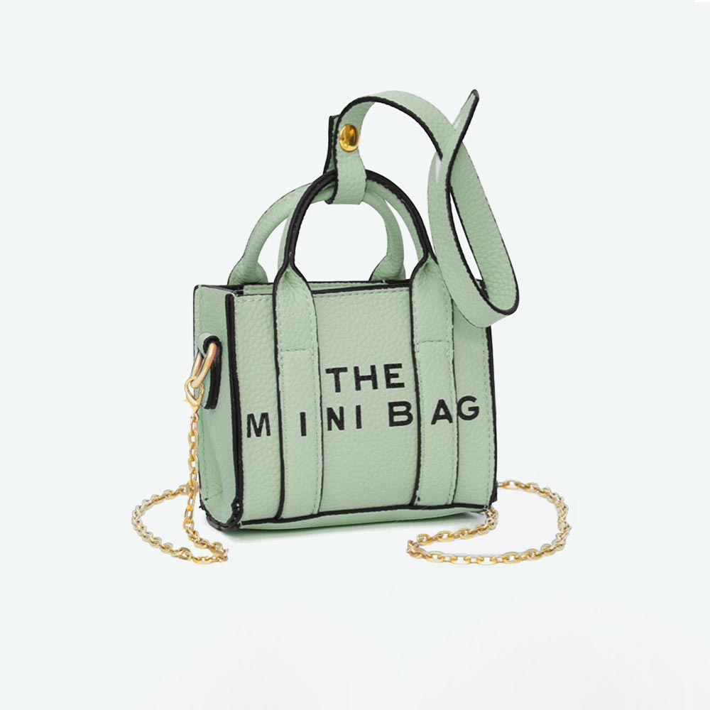 A mint green mini tote bag with black trim and the words 'THE MINI BAG' printed in black on the front. It features a mint green leather strap, a gold-tone chain strap, and a gold-tone metal snap closure.