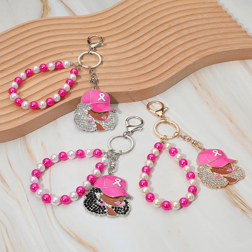 Three Afro woman awareness ribbon keychain bag charms displayed on wood and marble surface — silver-tone with clear rhinestones and pink cap (top left), silver-tone with black rhinestones and pink and white pearl wristlet (bottom center), gold-tone with clear rhinestones and pink and white pearl wristlet (bottom right), all featuring hot pink enamel baseball cap with white ribbon charm