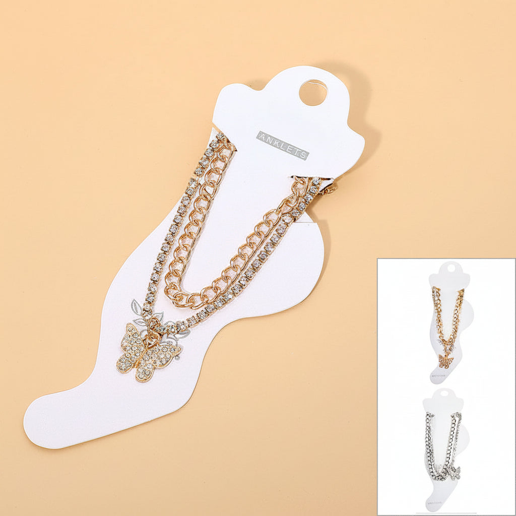 Rhinestone pavé butterfly charm layered chain anklet in gold-tone on white foot-shaped retail card, gold curb chain and rhinestone tennis chain layers with rhinestone pavé butterfly pendant, warm peach background, inset showing both gold-tone and silver-tone colorways on retail cards
