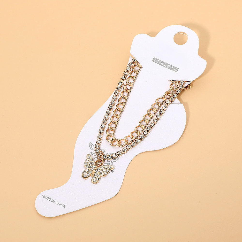 Rhinestone pavé butterfly charm layered chain anklet in gold-tone displayed on white foot-shaped retail card — gold curb chain and rhinestone tennis chain layers, rhinestone pavé butterfly pendant with leaf charm, gold lobster clasp, warm peach background