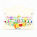 Kids charm beaded stretch bracelet set in pastel multicolor — 3-piece stackable set with pastel pearl bead and flower charm bracelet, colorful disc bead bracelet with smiley face accent, and LOVE charm bracelet with carrot, pineapple, heart, and letter charms, cream retail card, white background