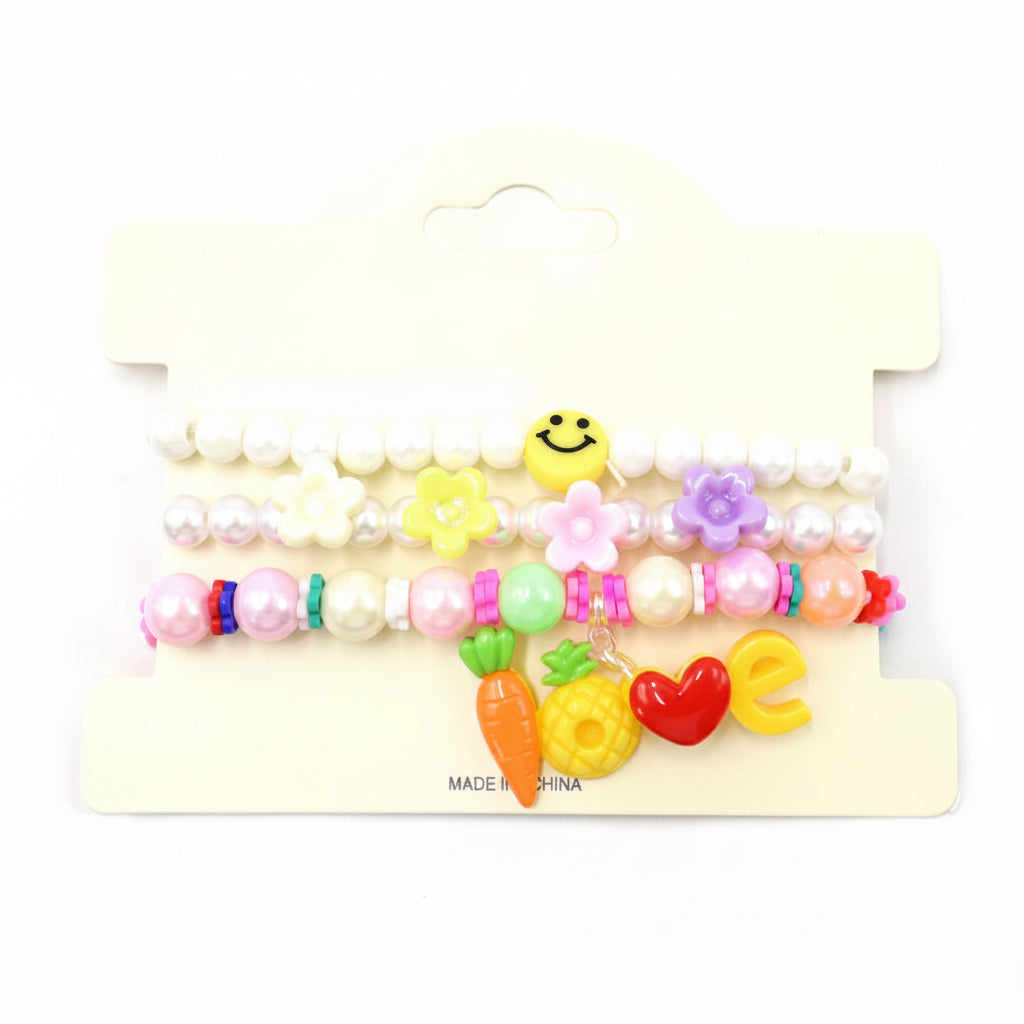Kids charm beaded stretch bracelet set in white pearl — 3-piece stackable set with white pearl bead bracelet with smiley face, flower charm bracelet with yellow and lavender flowers, and LOVE charm bracelet with carrot, pineapple, heart, and letter charms, cream retail card, white background