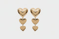 Triple metal heart link dropdown earrings in polished gold-tone — large puffy heart post stud at top with two cascading smaller puffy heart charms linked by gold jump rings, graduating in size from medium to small, warm gray background