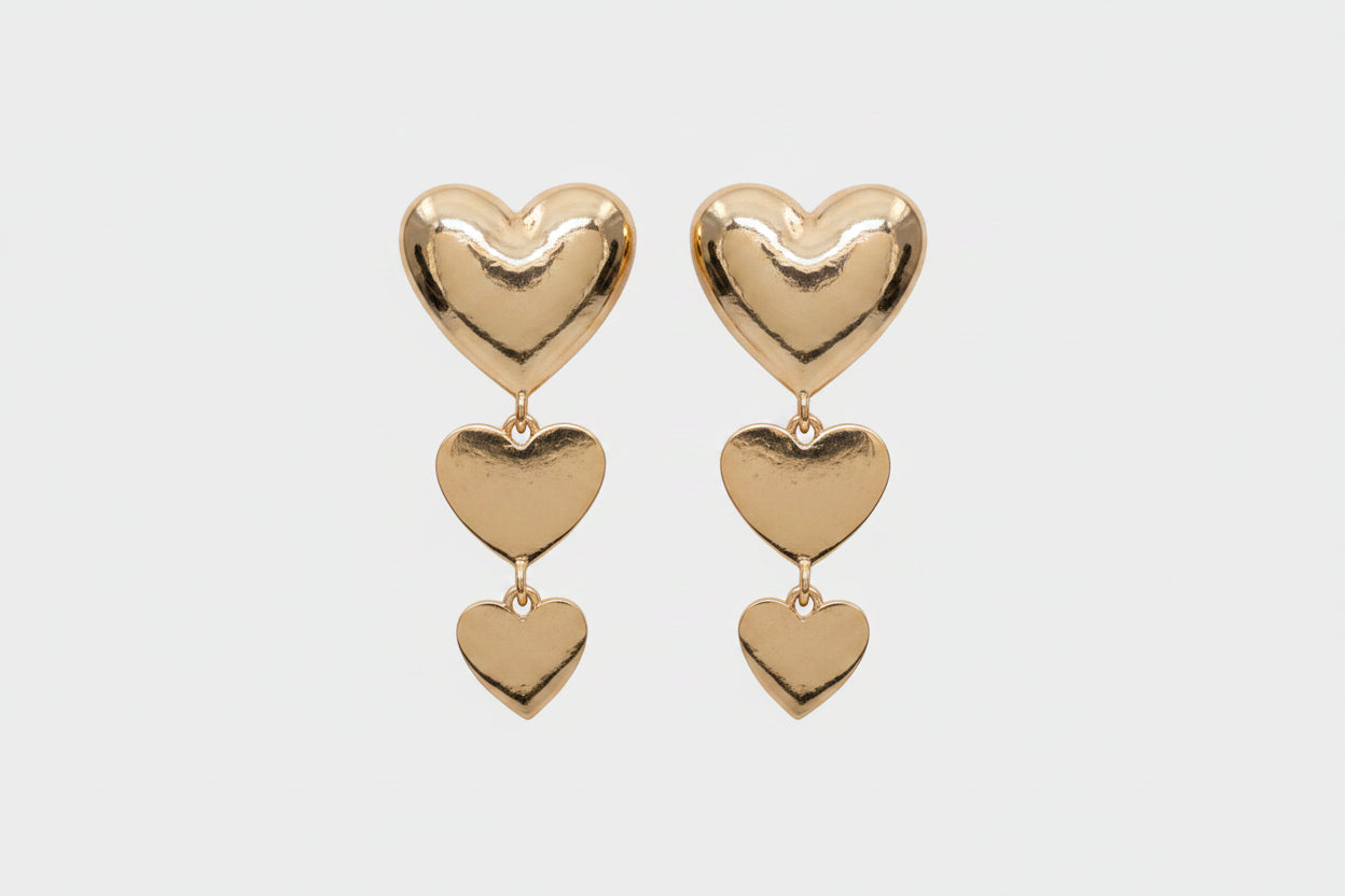 Triple metal heart link dropdown earrings in polished gold-tone — large puffy heart post stud at top with two cascading smaller puffy heart charms linked by gold jump rings, graduating in size from medium to small, warm gray background