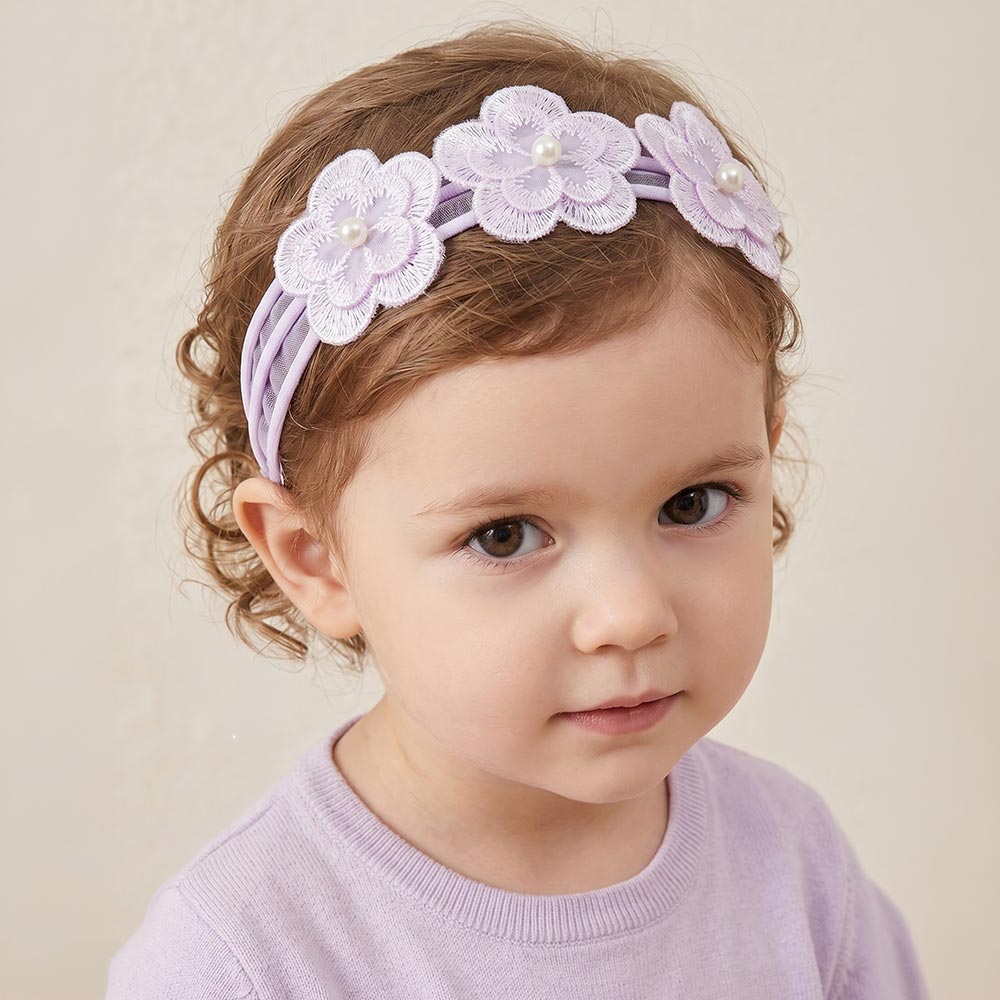 A toddler with curly brown hair wearing a lavender purple fabric headband decorated with three white embroidered flowers and small pearl-like centers, paired with a matching lavender top against a soft beige background.