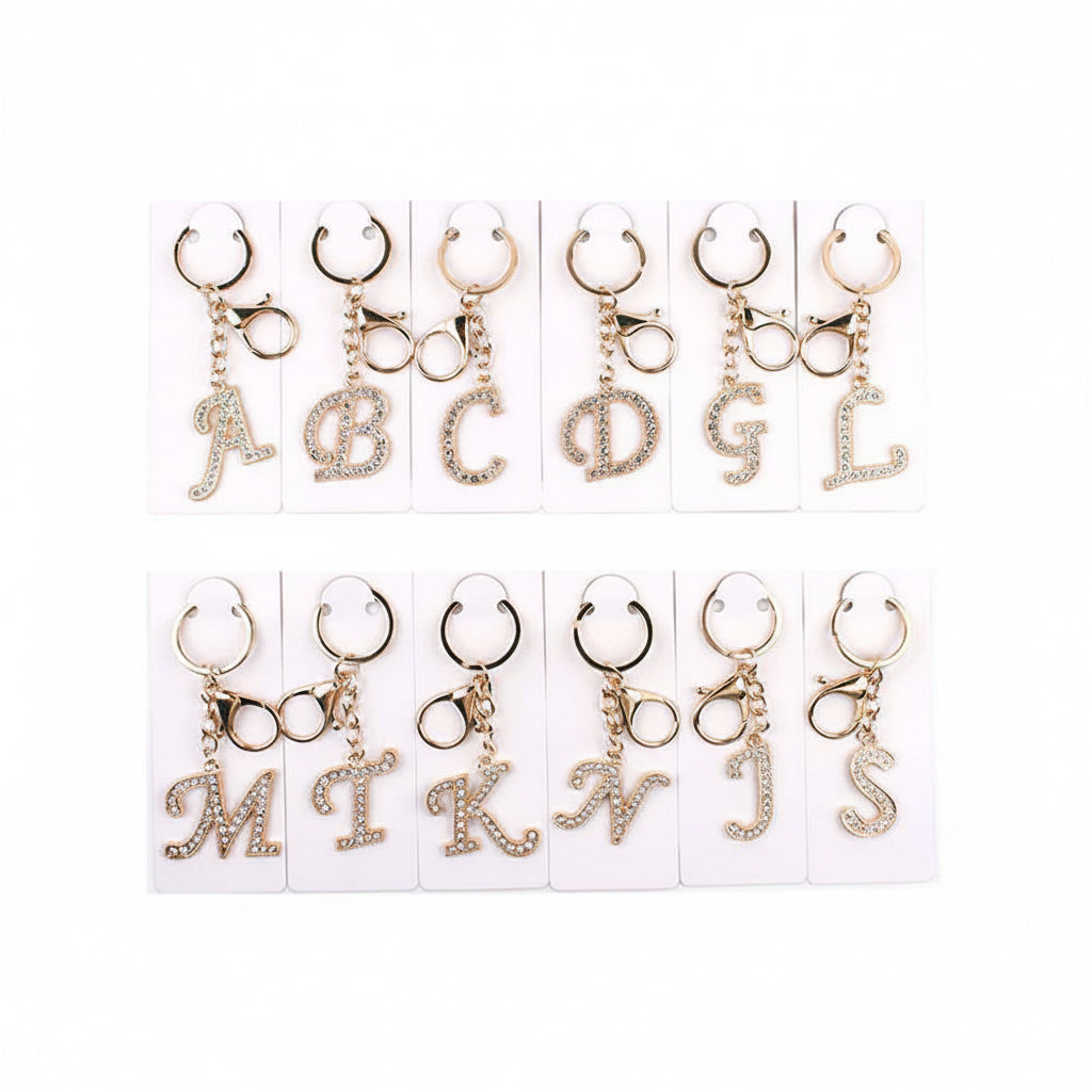 12-piece assorted rhinestone pavé initial letter keychain bag charm set displayed on white retail cards — letters A, B, C, D, G, L, M, T, K, N, J, S — clear crystal rhinestone pavé script letter pendants, gold-tone lobster clasps and key rings, white background