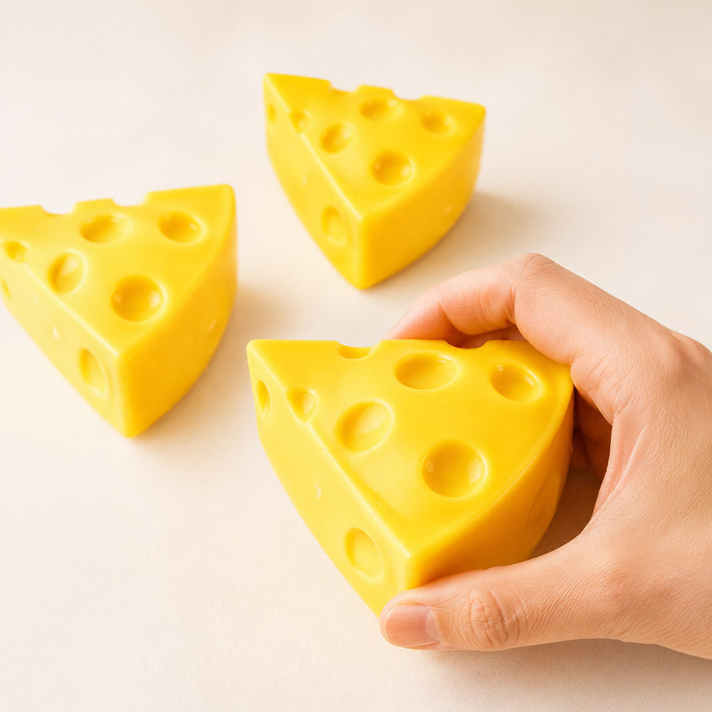 Hand squeezing yellow cheese wedge squishy stress relief toy showing soft foam deformation, two additional cheese wedge squishies in background on cream surface
