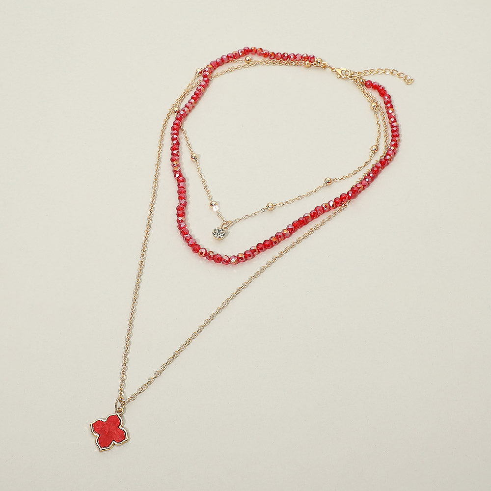 Red faceted crystal beaded layered necklace with gold satellite chain and red quatrefoil clover pendant — flat lay on cream background showing rhinestone accent charm