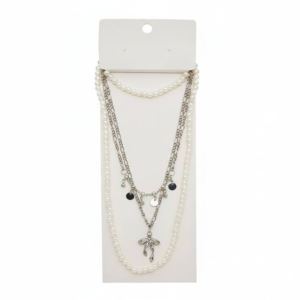 Silver-tone pearl bow pendant layered chain necklace on white display card — faux pearl strand, silver figaro chain with disc charms, and sculptural silver bow pendant, white background