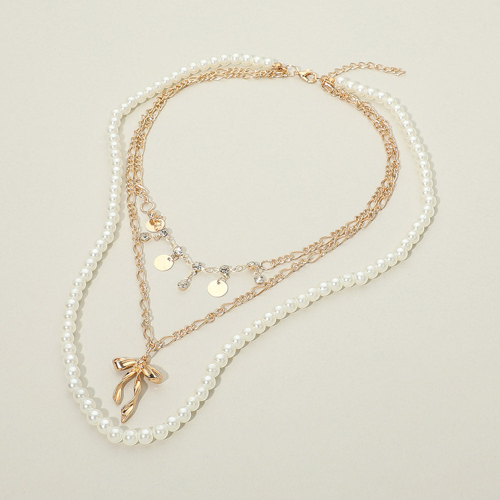 Gold-tone pearl bow pendant layered necklace flat lay on cream background — faux pearl beaded strand, gold figaro chain with rhinestone accents and flat disc charms, sculptural gold bow pendant