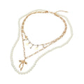 Gold-tone pearl bow pendant layered necklace flat lay on white background — faux pearl strand, gold figaro chain with rhinestone and disc charms, and 3D gold bow pendant with lobster claw clasp and extender chain