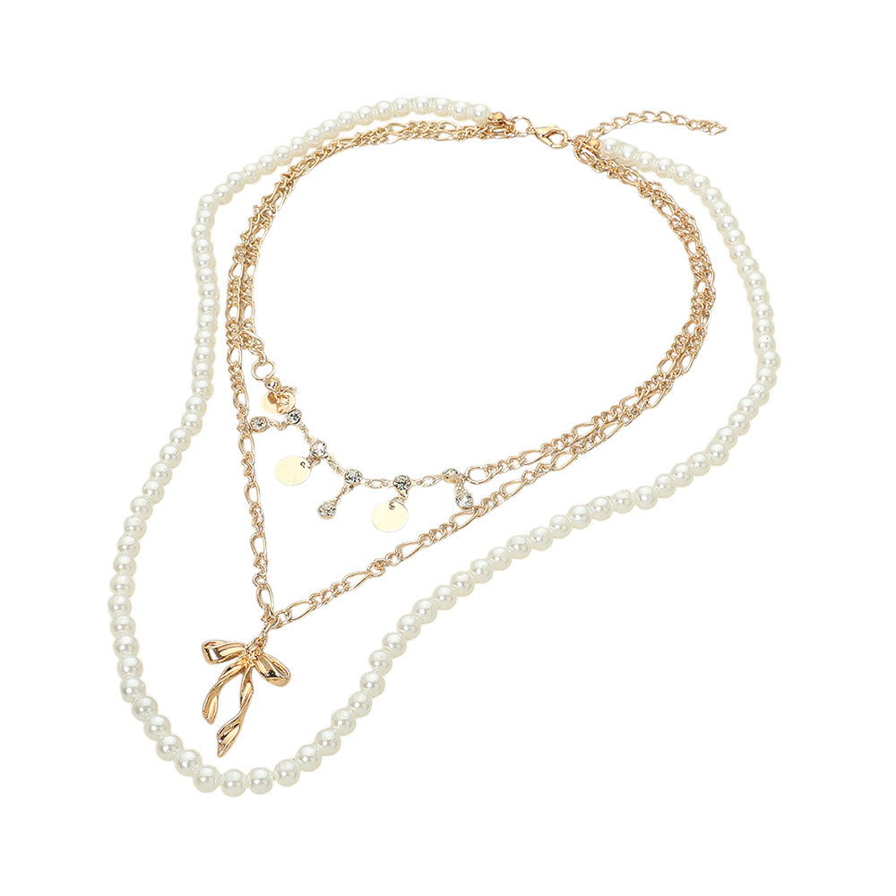 Gold-tone pearl bow pendant layered necklace flat lay on white background — faux pearl strand, gold figaro chain with rhinestone and disc charms, and 3D gold bow pendant with lobster claw clasp and extender chain