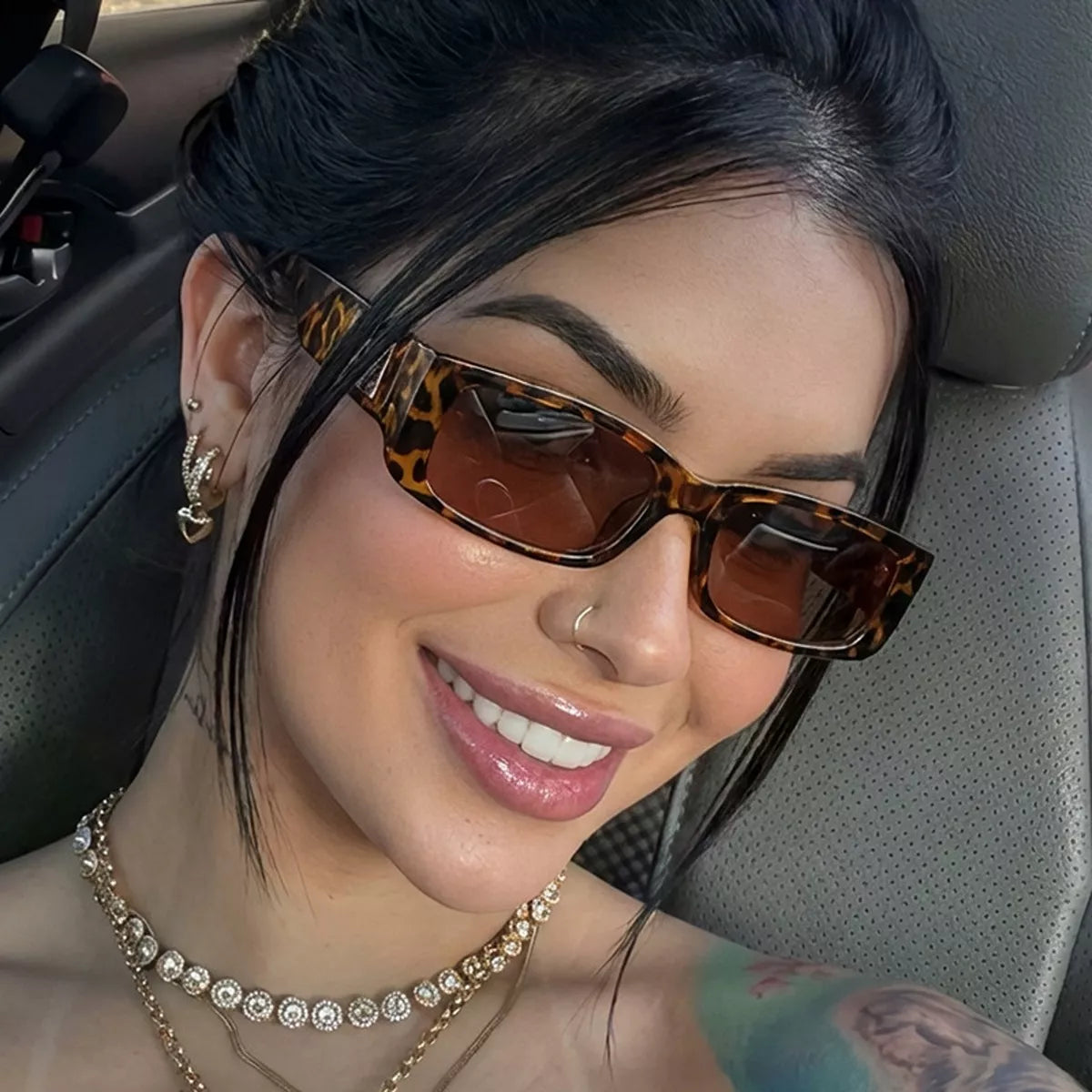 Smiling woman wearing brown tortoiseshell-pattern rectangular sunglasses with brown gradient lenses and layered necklaces inside a car