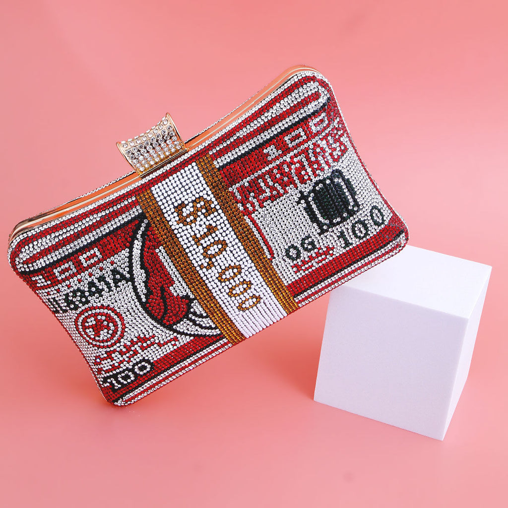 Women's red rhinestone cash clutch bag with 100 dollar bill crystal design, red and silver rhinestone embellishment with gold-tone stripe, crystal frame clasp, displayed upright next to white cube prop on pink background
