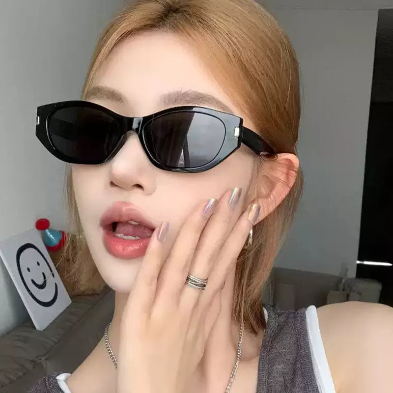 Woman with blonde hair wearing black angular cat-eye sunglasses with dark lenses and silver-tone corner accents, resting her hand against her cheek indoors