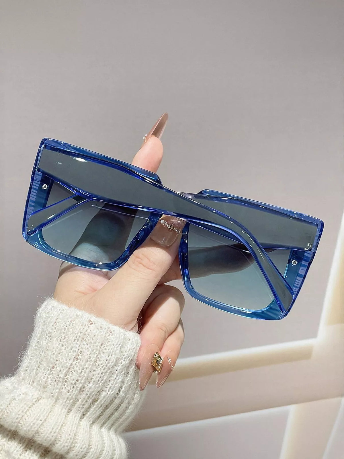 Rear view of blue square sunglasses with temples folded across the gradient lenses, held against a gray wall