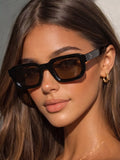 Model wearing glossy black square sunglasses with brown-tinted lenses and metallic-looking temple dots, styled with gold-tone earrings