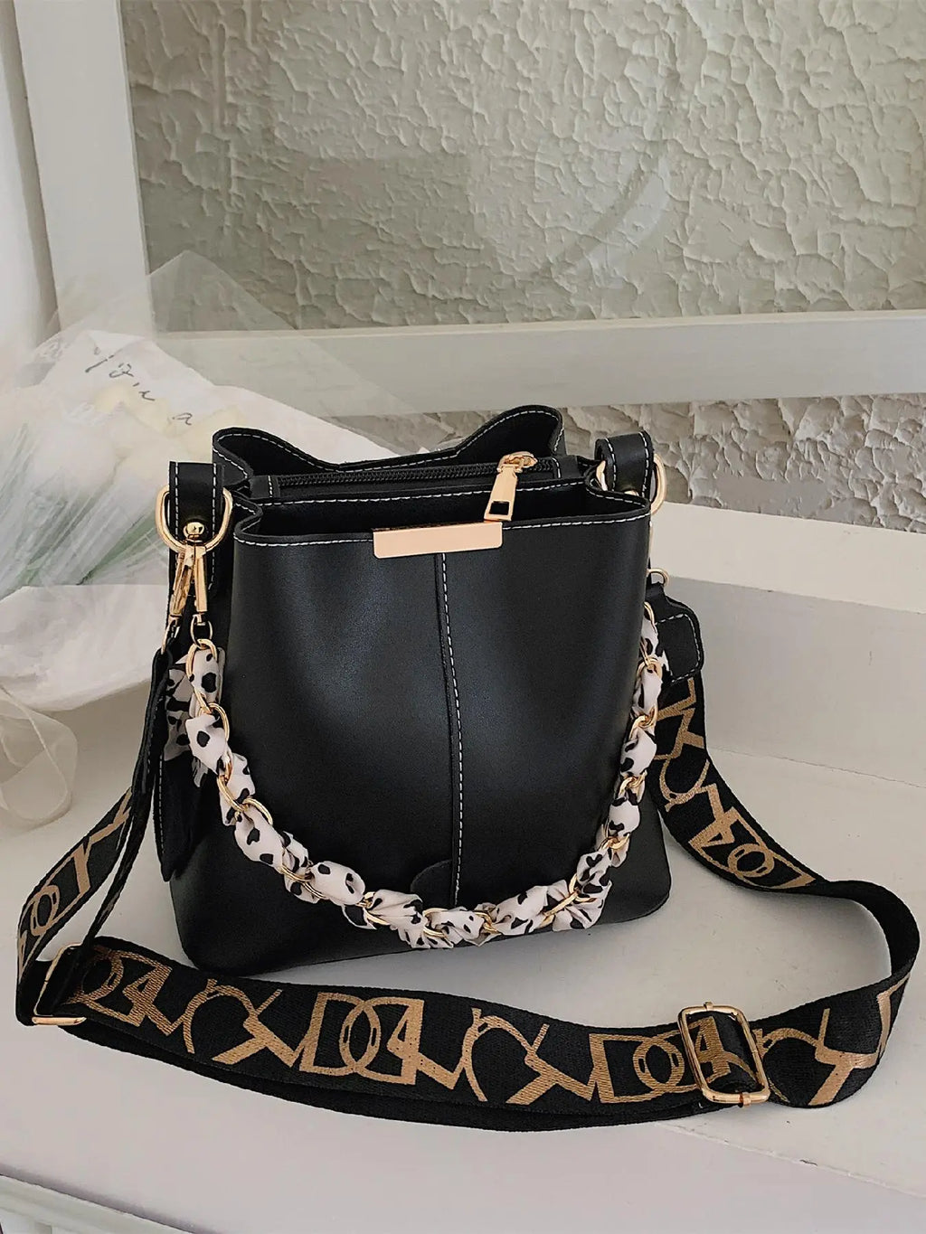 Black PU leather bucket crossbody bag with white contrast stitching, gold-tone chain and black-and-white leopard print silk scarf handle, black and gold letter-print wide crossbody strap, gold rectangular hardware plate and gold zipper pull, styled on white shelf with textured wall background
