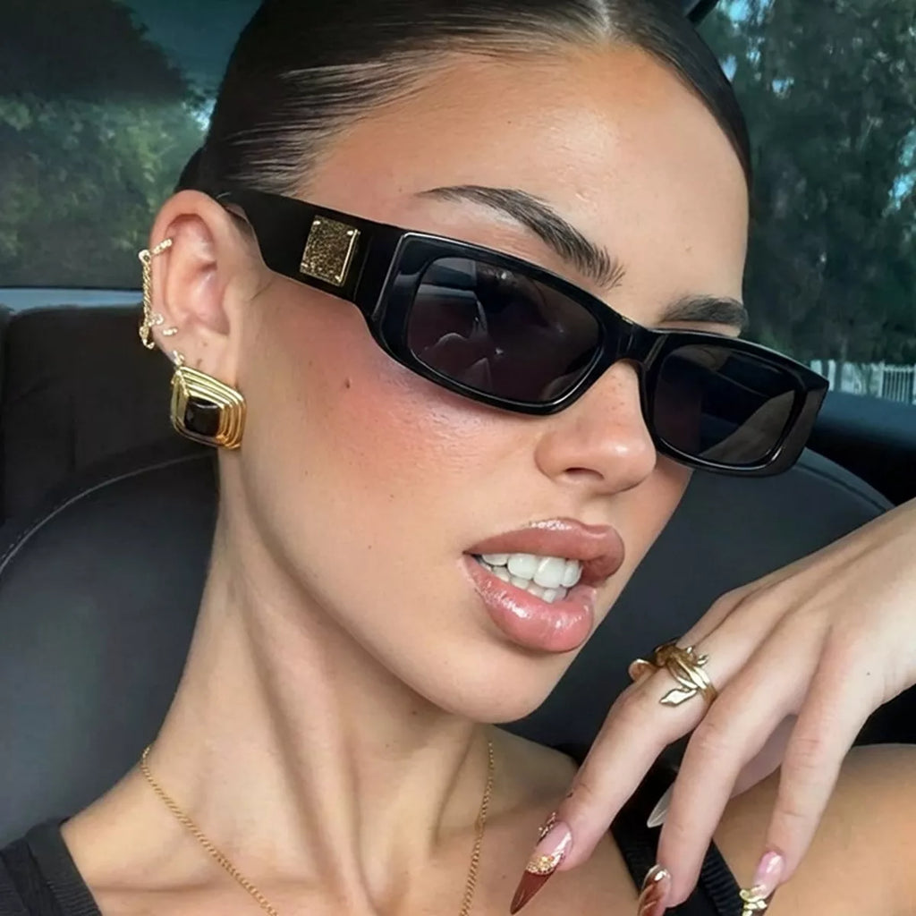Woman wearing black rectangular sunglasses with dark lenses and gold-tone temple accents, posing with her hand near her face inside a car