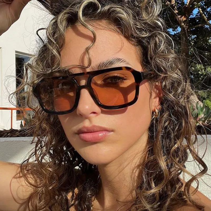 Woman with highlighted curly hair wearing tortoiseshell-pattern square sunglasses with amber-tinted lenses outdoors near a white wall and trees