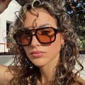 Woman with highlighted curly hair wearing tortoiseshell-pattern square sunglasses with amber-tinted lenses outdoors near a white wall and trees