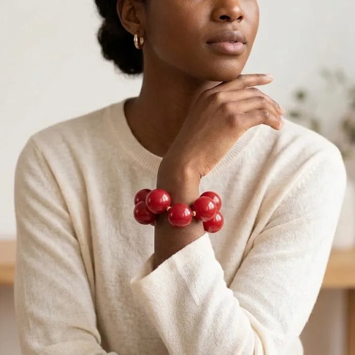 Model wearing a red bubble bead bracelet with a cream sweater, her hand raised near her chin in a softly blurred indoor setting