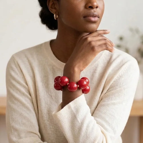 Model wearing a red bubble bead bracelet with a cream sweater, her hand raised near her chin in a softly blurred indoor setting