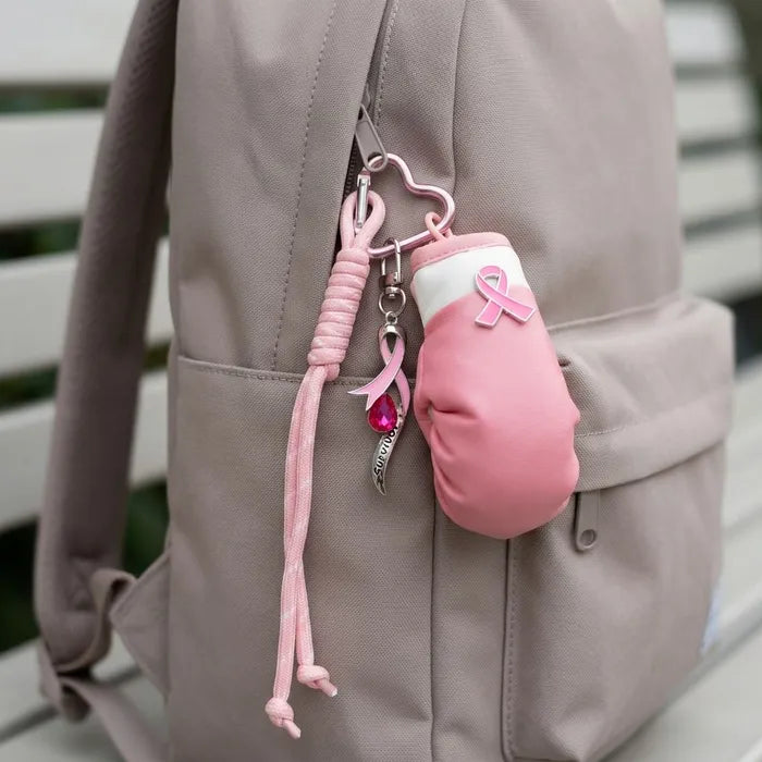 A pink breast cancer awareness themed keychain bag charm hanging from a taupe-gray backpack. The charm features a pink boxing glove with a white top section, a pink ribbon, a silver heart-shaped clasp, and a pink cord.