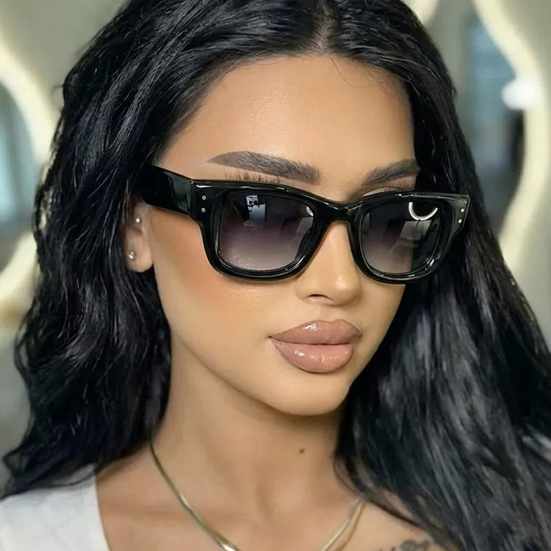Woman with long black hair wearing glossy black oversized square sunglasses with smoky gradient lenses and paired metallic corner details, styled with a white top and necklace