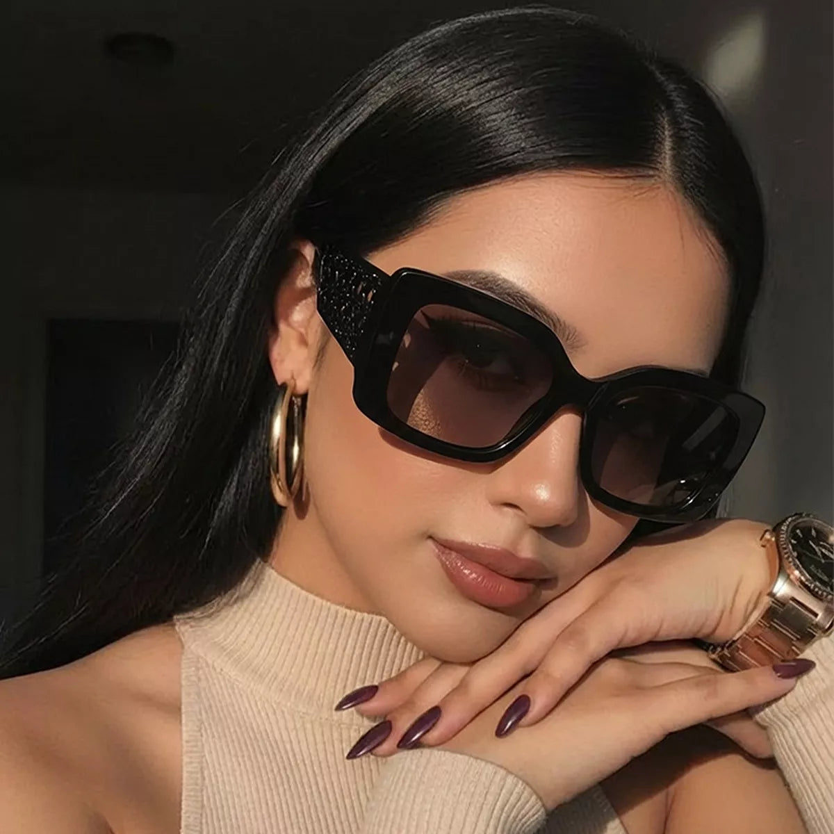 Model wearing black oversized square sunglasses with brown gradient lenses and wide lattice temples, a beige knit top, gold hoop earrings and a gold-tone watch, resting her chin on her hands
