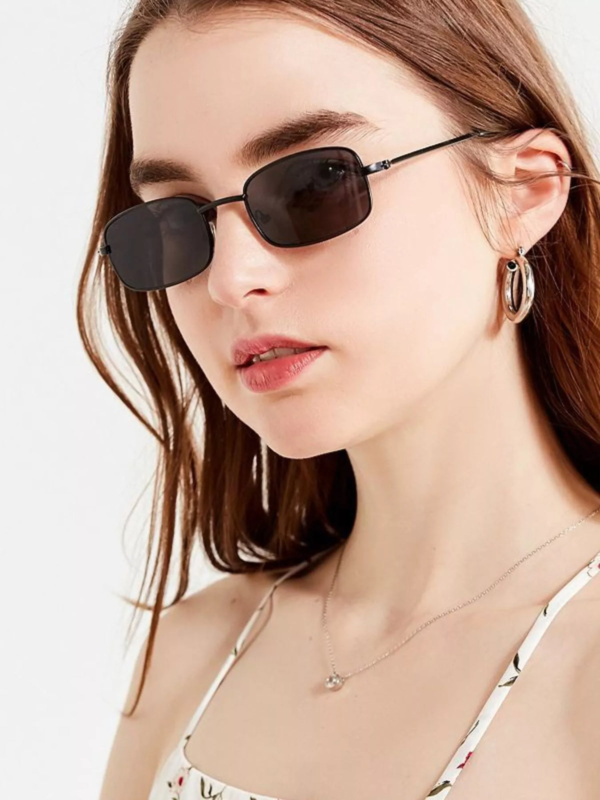Woman with straight brown hair wearing slim black rectangular metal sunglasses with dark lenses, hoop earrings, and a floral spaghetti-strap top against a white background