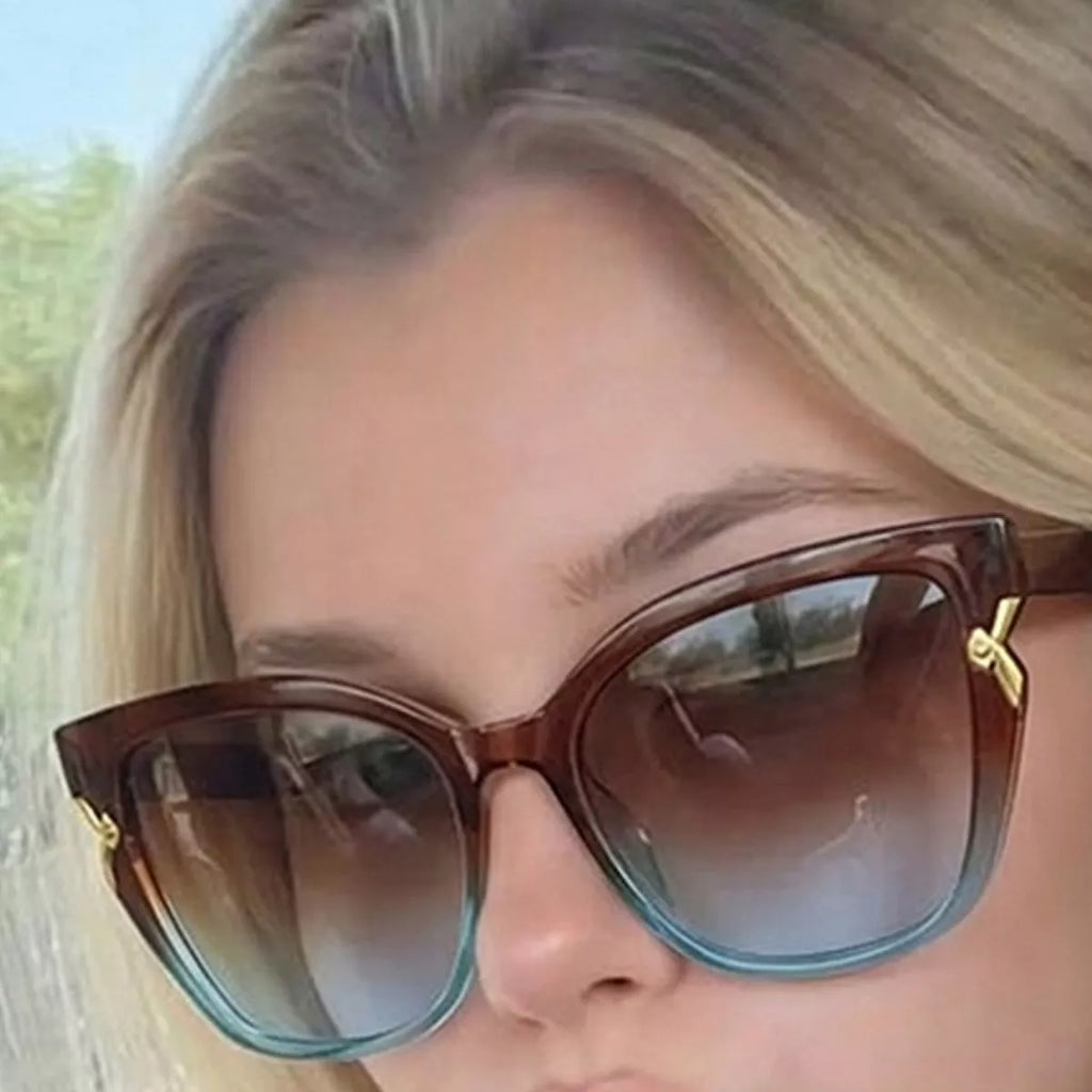 Close-up of brown-to-teal cat-eye sunglasses with brown-to-blue-gray gradient lenses and gold-tone corner accents on a blonde woman