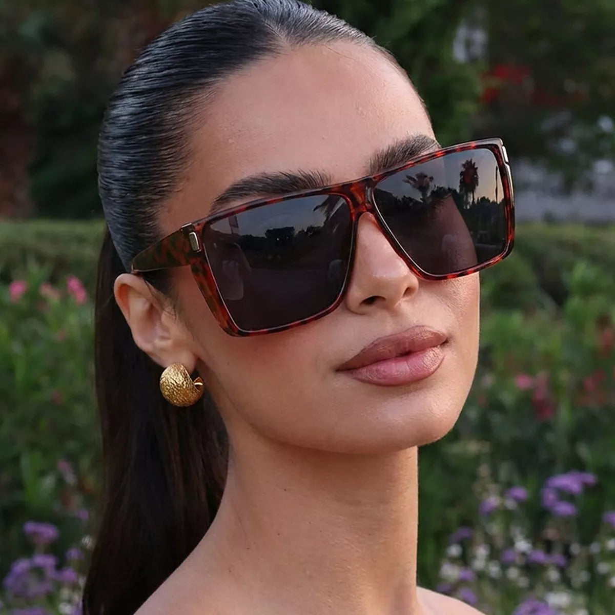 Model wearing oversized tortoiseshell-pattern square sunglasses with dark lenses and gold-tone earrings in a garden