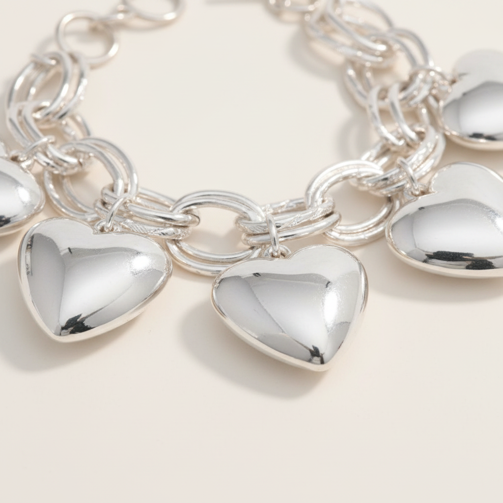 Close-up macro of chunky silver-tone double oval link chain bracelet with three large polished puffy hollow heart charms dangling, high-gloss reflective finish, warm cream background with soft shadow