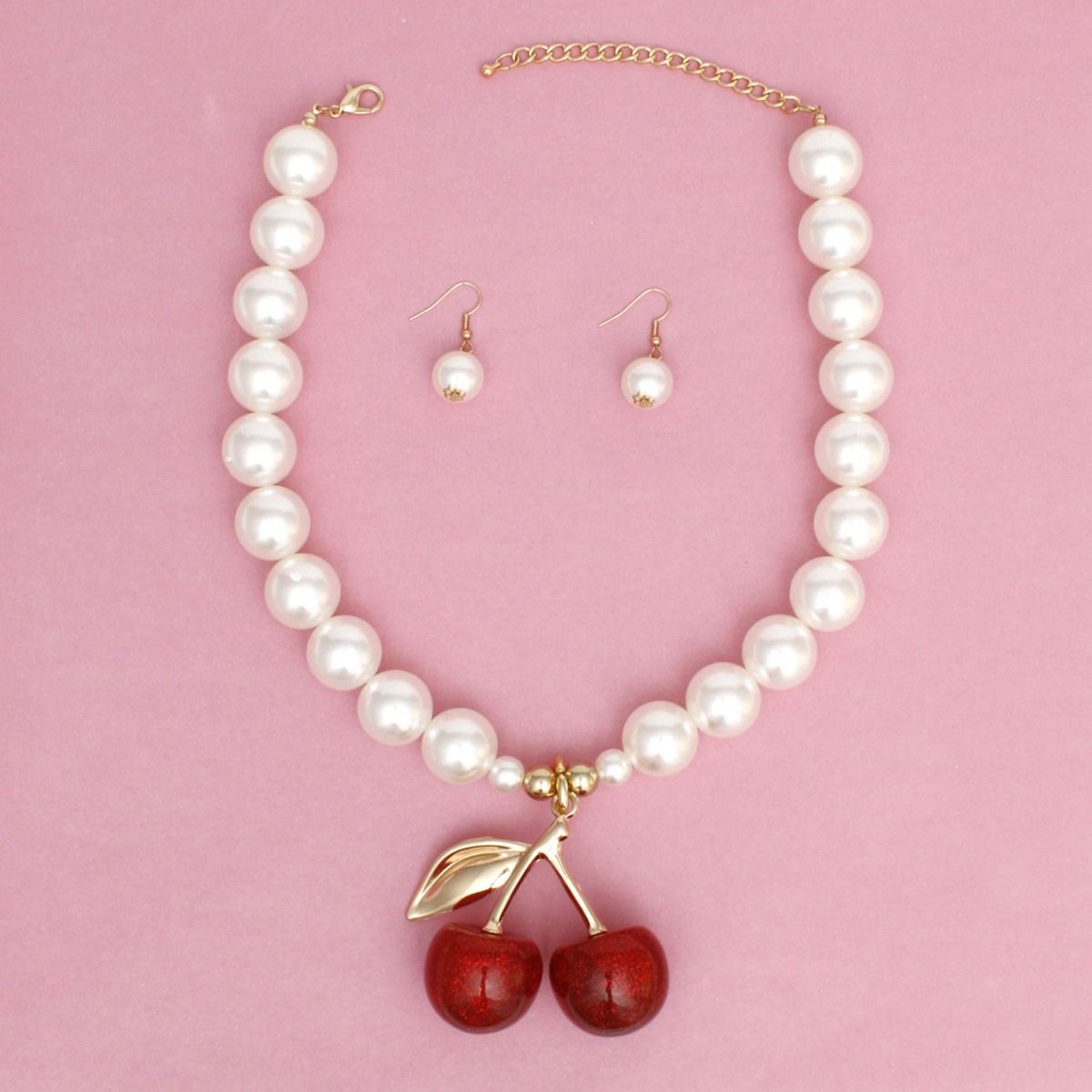 Cream faux pearl necklace with a red double-cherry pendant, gold-tone stem and leaf, lobster clasp and extender, and matching drop earrings on a pink background