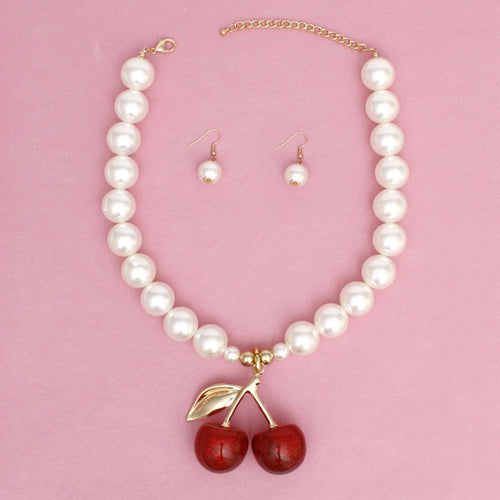 Cream faux pearl necklace with a red double-cherry pendant, gold-tone stem and leaf, lobster clasp and extender, and matching drop earrings on a pink background