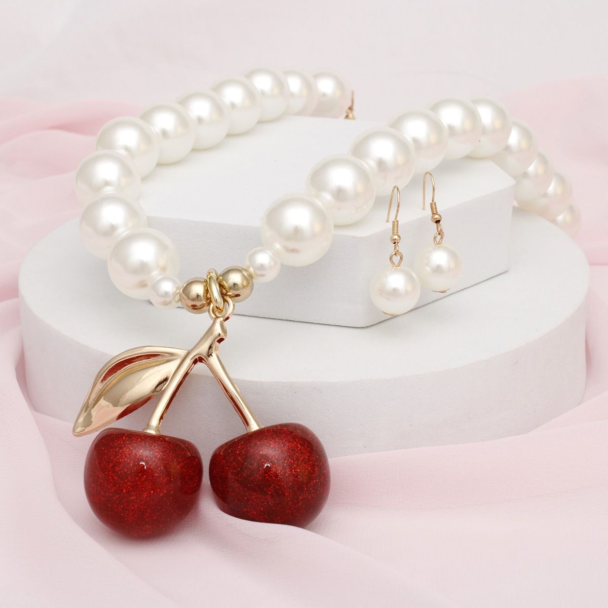 Close-up of red glitter cherries with a gold-tone stem and leaf on a cream faux pearl necklace, displayed with matching earrings on white blocks and pink fabric