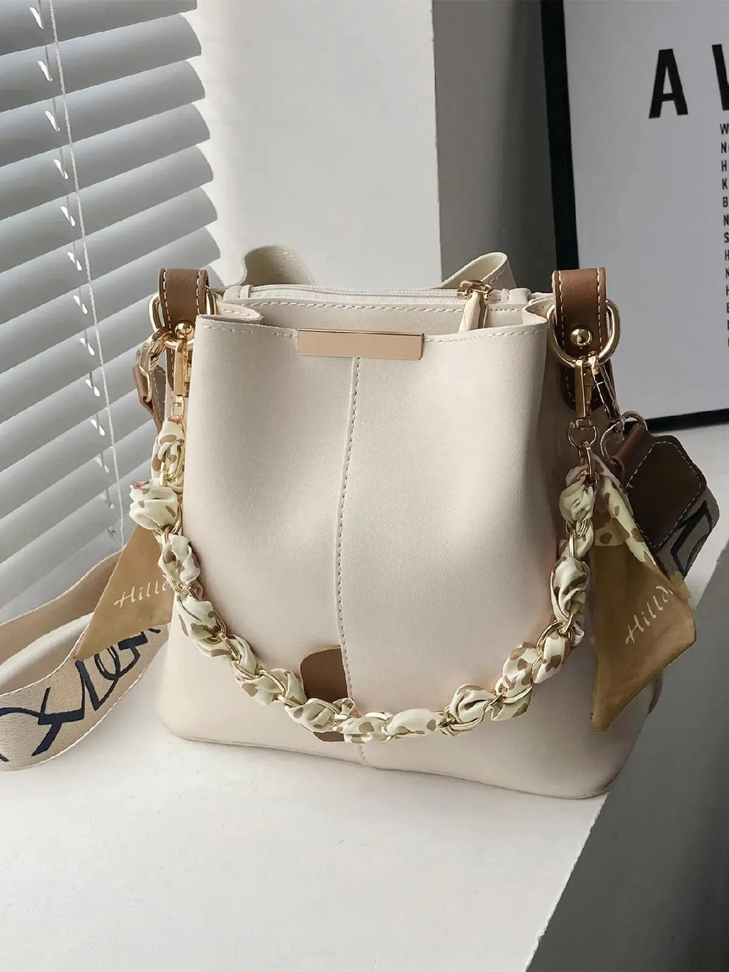 Cream beige PU leather bucket crossbody bag with tan leather trim accents, gold-tone chain and cream leopard print silk scarf handle, beige and black letter-print wide crossbody strap with gold buckle, gold rectangular hardware plate, styled on white shelf with window blinds and framed art print in background