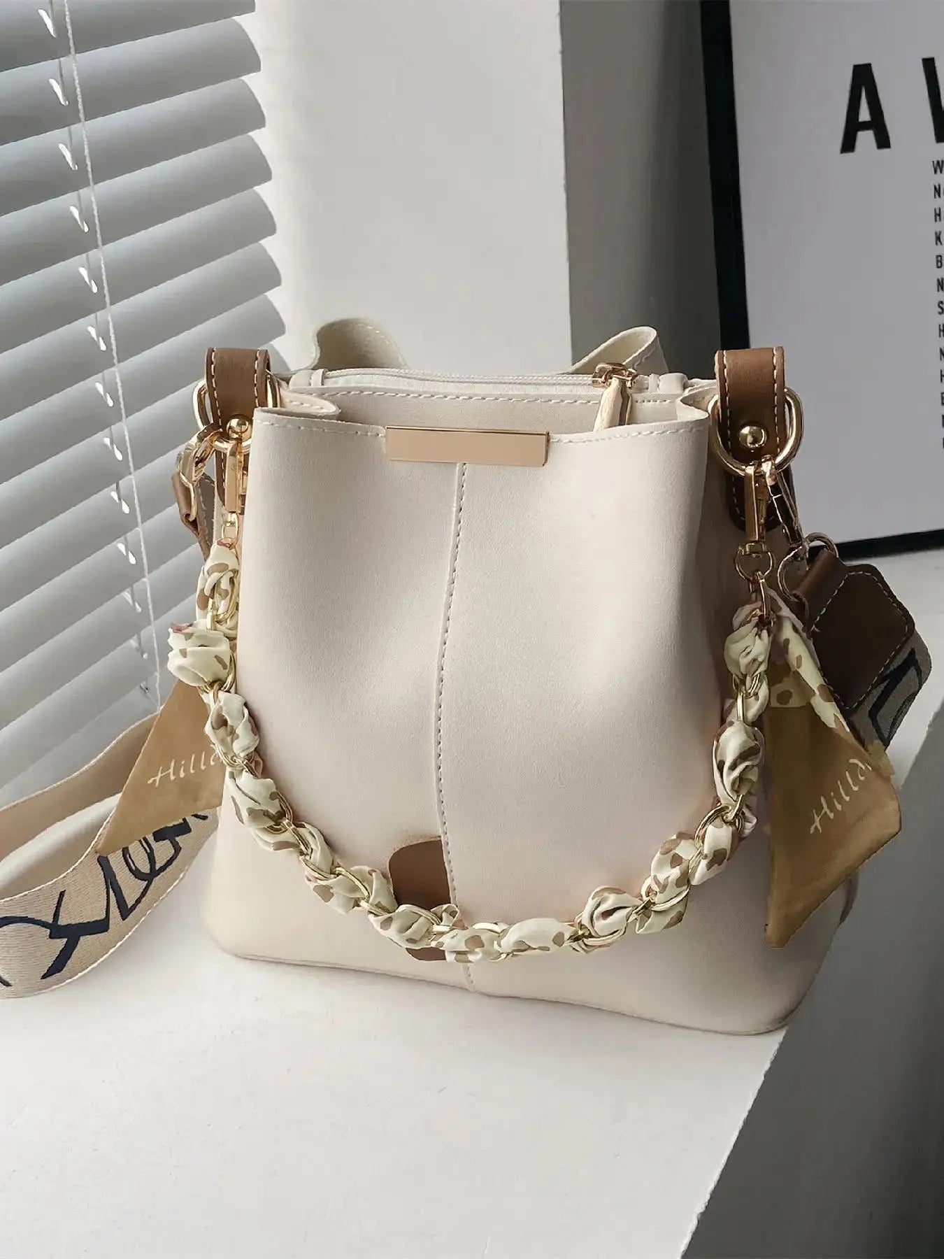 Cream beige PU leather bucket crossbody bag with tan leather trim accents, gold-tone chain and cream leopard print silk scarf handle, beige and black letter-print wide crossbody strap with gold buckle, gold rectangular hardware plate, styled on white shelf with window blinds and framed art print in background