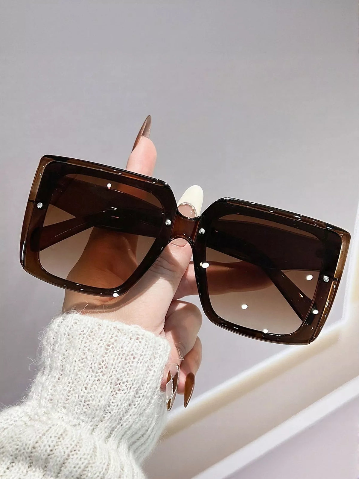 Coffee-brown oversized square sunglasses with brown gradient lenses, held by a manicured hand in a white knit sleeve against a gray wall