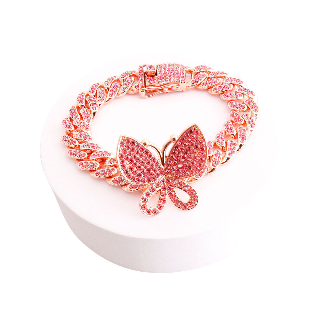Overhead angled shot of a fully pave pink rhinestone Miami Cuban chain link bracelet in rose gold tone arranged in a circle on a round white cylindrical display stand against a white background. An oversized fully pave pink rhinestone butterfly charm measuring 1.25 inches is centered at the bottom. A rhinestone-crusted square push-lock box clasp with a hinged safety bar is visible at the top.