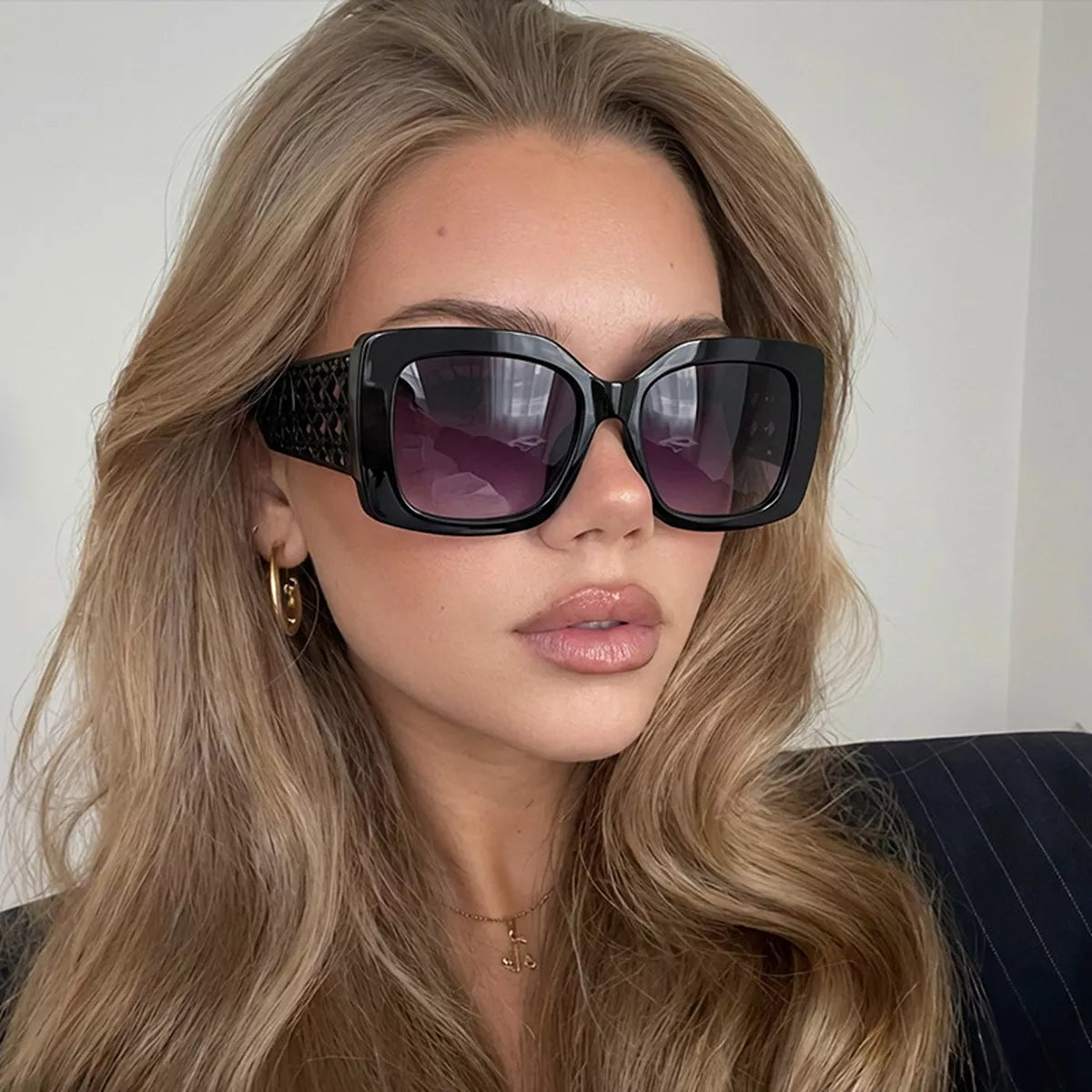 Model wearing black oversized square sunglasses with purple gradient lenses and wide lattice temples, gold hoop earrings, a delicate necklace and a dark pinstriped outfit against a light background
