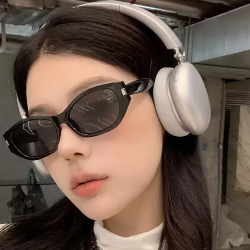 Angled close-up of a woman wearing black cat-eye sunglasses with dark lenses, silver-tone corner accents, and silver over-ear headphones in an industrial-style interior