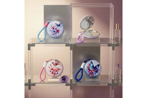 Four round hard-shell butterfly floral compact mirror keychains displayed in individual clear acrylic cube shelves. Top left shows a white case with blue and pink butterfly floral print and blue rope wrist strap with pink wrapped section. Top right shows a lavender case open revealing two oval silver mirror panels with lavender rope loop. Bottom left shows a white case with orange and pink butterfly floral print and pink rope wrist strap. Bottom right shows a light blue case with deep blue butterfly and pur