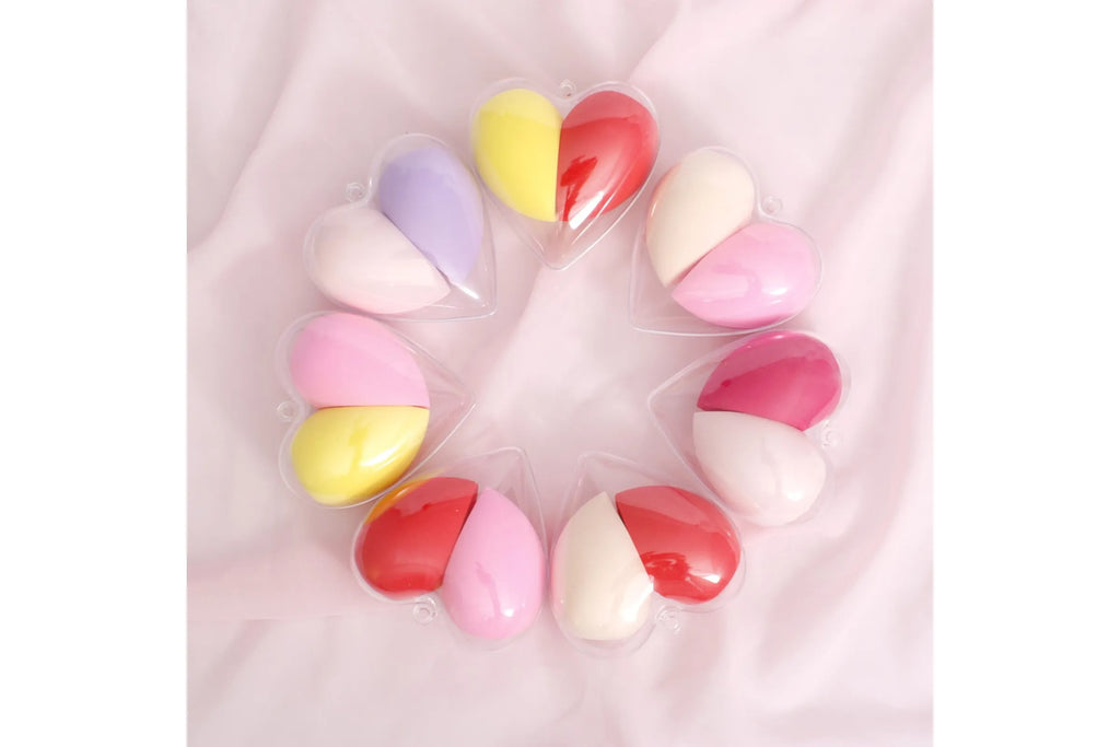 Assorted heart-shaped clear plastic cases with dual makeup beauty blenders in multiple color combinations arranged in a circle
