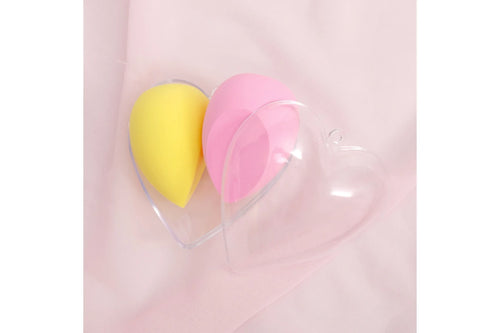 Close-up overhead view of a clear heart-shaped hard plastic case containing one yellow teardrop foam makeup sponge on the left and one pink teardrop foam makeup sponge on the right, resting on a soft blush pink fabric background.